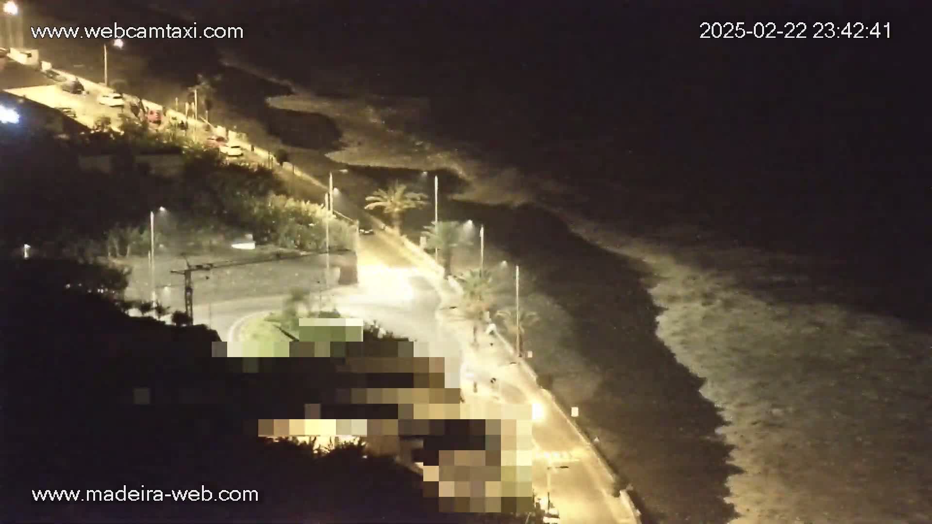 A nighttime, high-angle view shows a coastal road with some cars and pedestrians, alongside a dark ocean with waves crashing on the shore under artificial light.