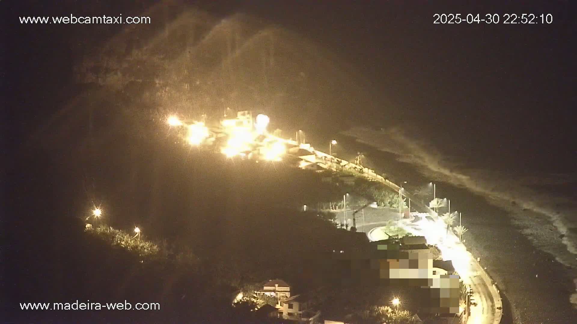 A nighttime coastal scene shows brightly lit buildings and a road curving along a dark beach under somewhat overcast conditions.