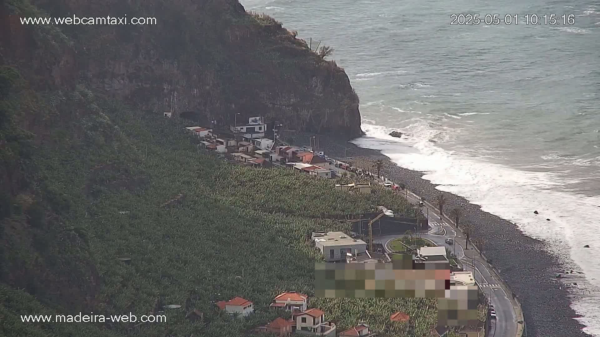 A coastal scene shows a dark-colored beach with waves crashing against the shore, next to a road that runs along a cliffside lined with banana plantations and small buildings, under overcast skies.