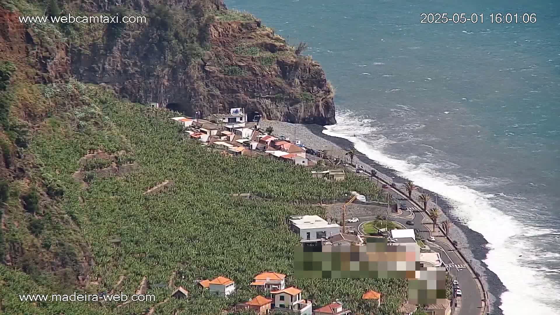 A coastal village nestled beside a dark-sand beach with moderate waves under a mostly sunny sky is surrounded by lush green banana plantations and a rocky cliff.