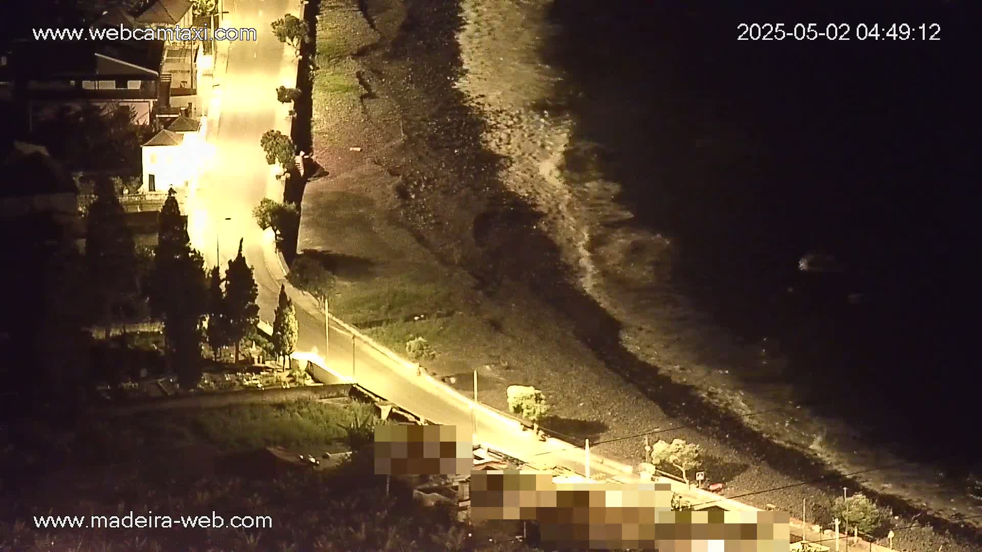 A nighttime, overhead view shows a paved road curving alongside a dark, rocky shoreline with small waves lapping against it, illuminated by streetlights.