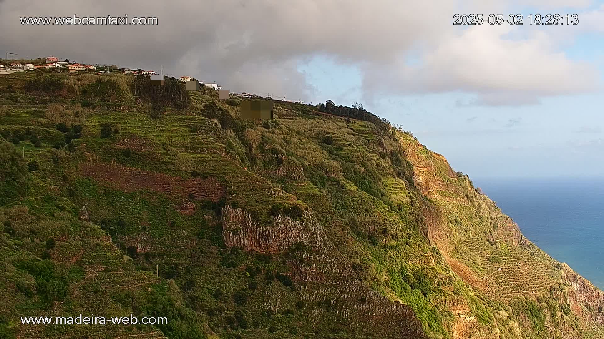 A verdant, terraced hillside descends to a blue ocean under a partly cloudy sky.