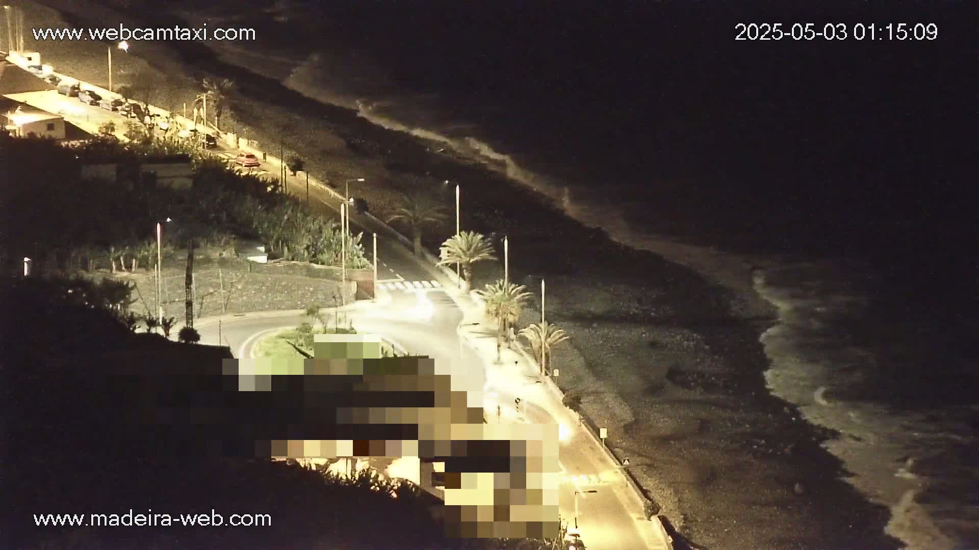A nighttime coastal road with palm trees curves alongside a dark beach with moderate waves under a clear, dark sky.