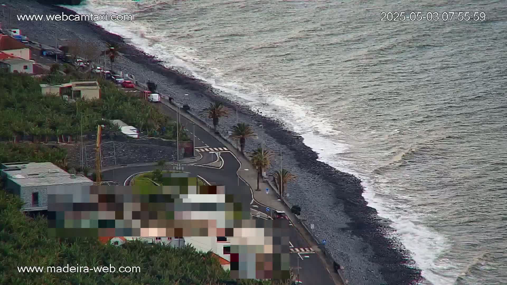A dark-grey, pebble beach with moderate waves laps against the shore next to a road lined with palm trees and buildings.