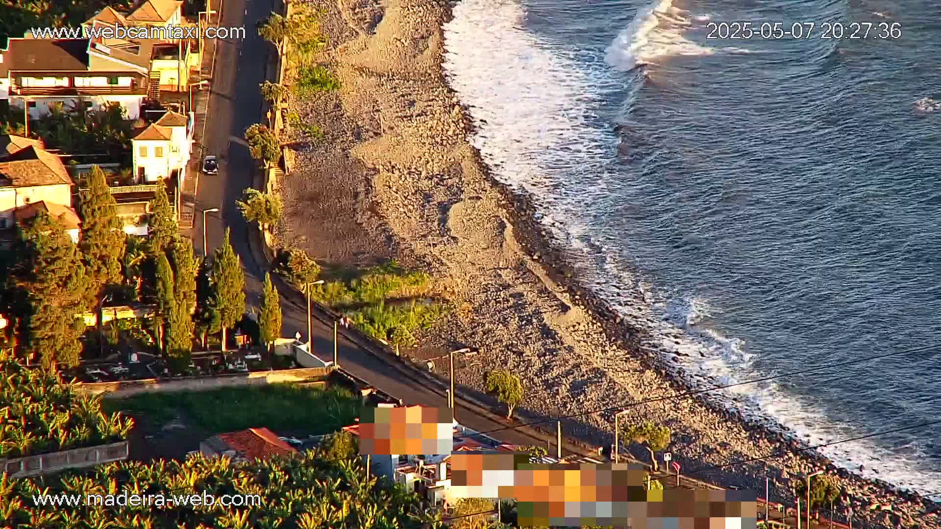 An aerial view shows a rocky coastline with gentle waves lapping the shore next to a road lined with houses and trees, under what appears to be a clear evening sky.