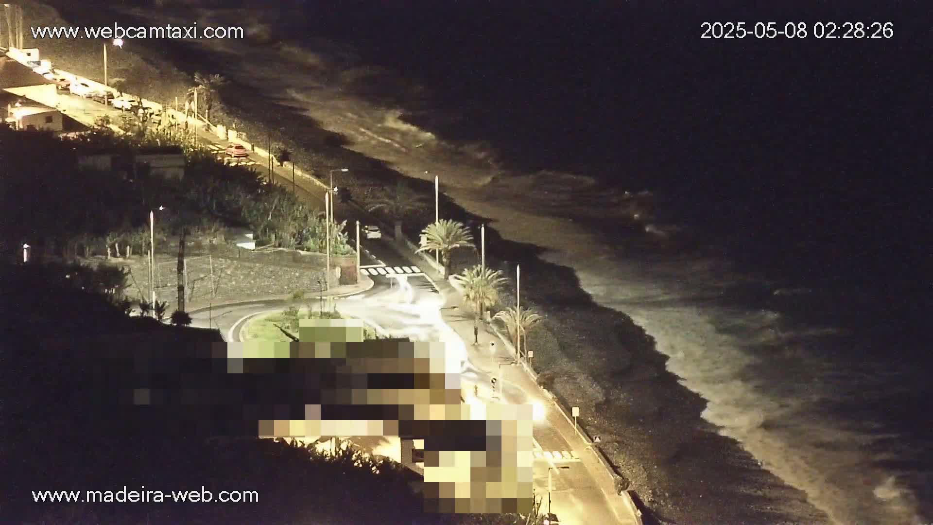 A nighttime coastal road scene shows waves crashing on a dark beach next to palm trees and buildings under artificial light.