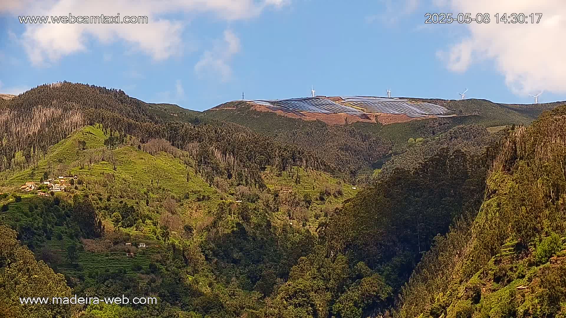 A mountaintop covered in solar panels and wind turbines is visible above a lush, green, hilly landscape under a partly cloudy blue sky.