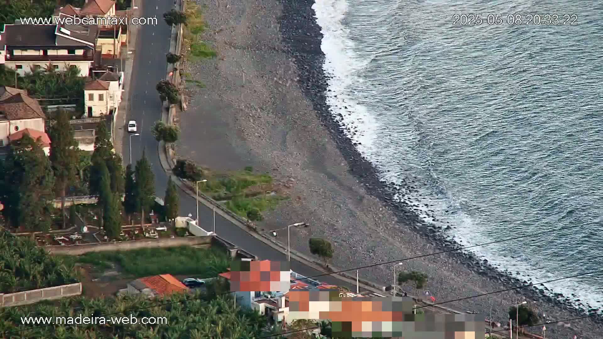 An aerial view shows a rocky coastline with gentle waves, a road winding alongside it past buildings and a small cemetery.