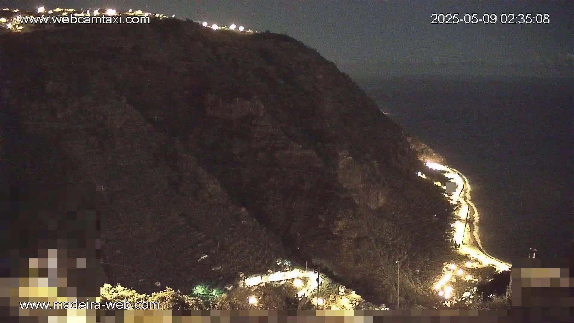 A dark, nighttime view shows a cliffside road illuminated by lights winding down towards a dark body of water under a clear night sky.