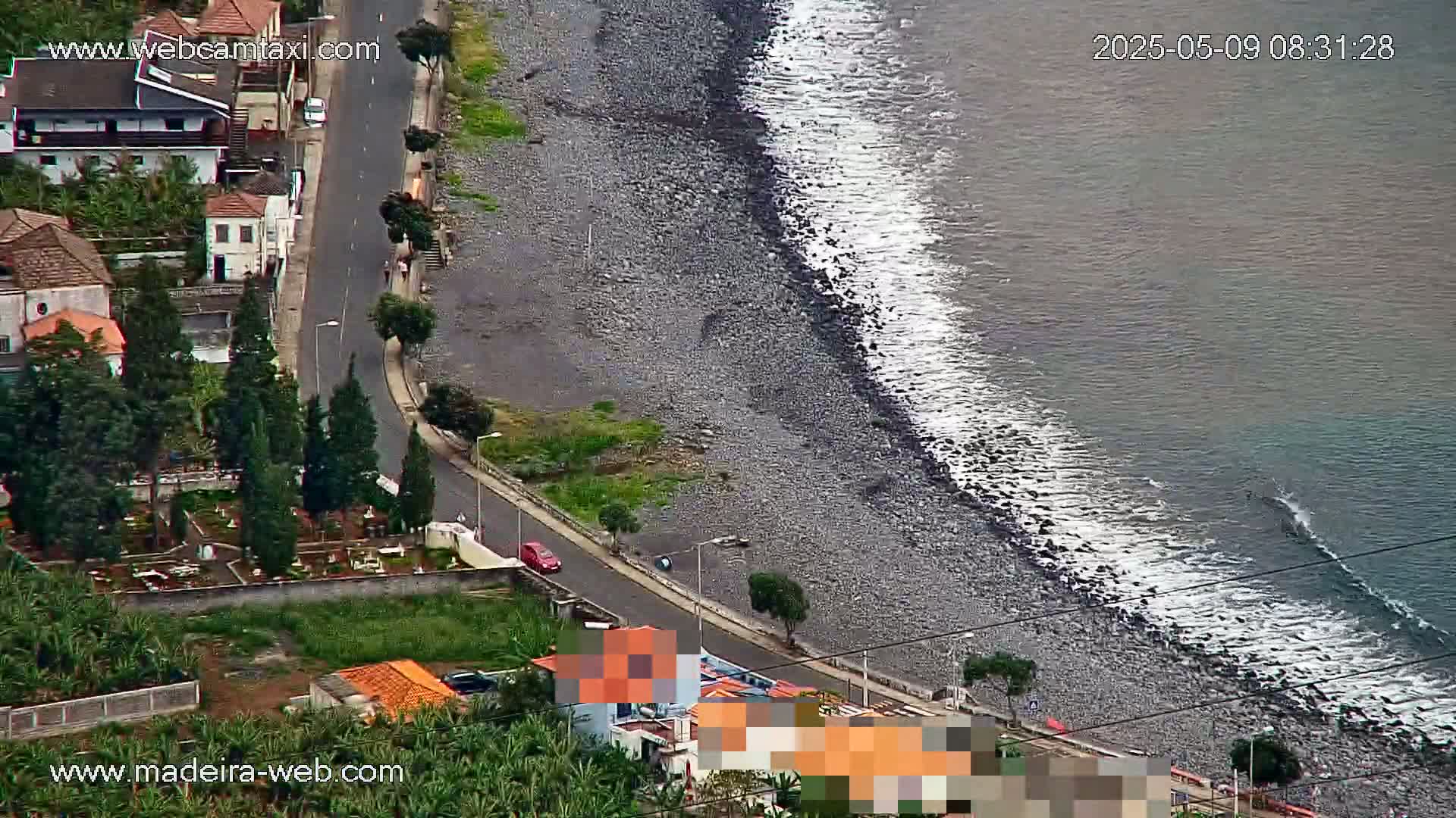 An aerial view shows a rocky beach with small waves lapping the shore next to a coastal road lined with houses and vegetation.