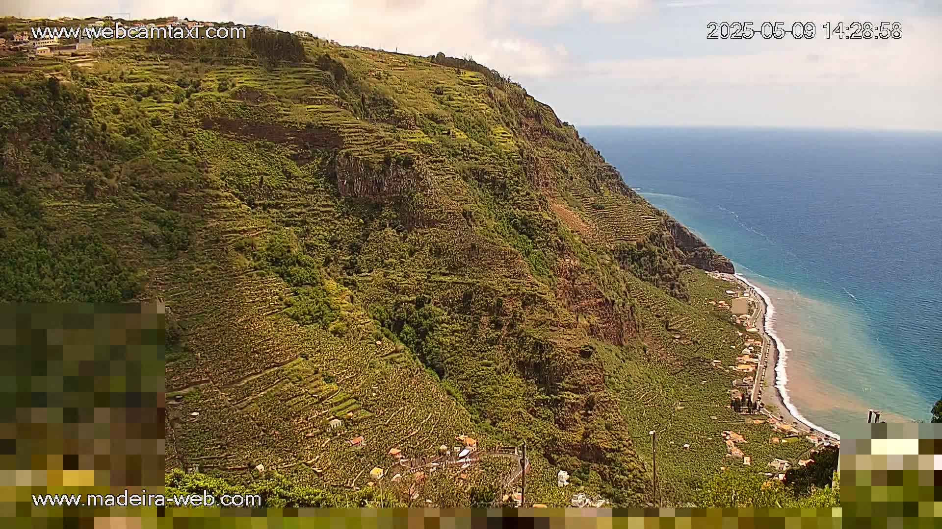 A steep, terraced hillside covered in vegetation slopes down to a dark-sand beach and calm ocean under a partly cloudy sky.