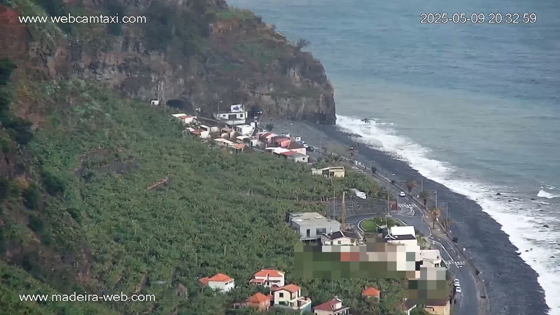 A coastal village nestled beside a dark-sand beach with moderate waves under an overcast sky, surrounded by lush green vegetation on a steep cliffside.