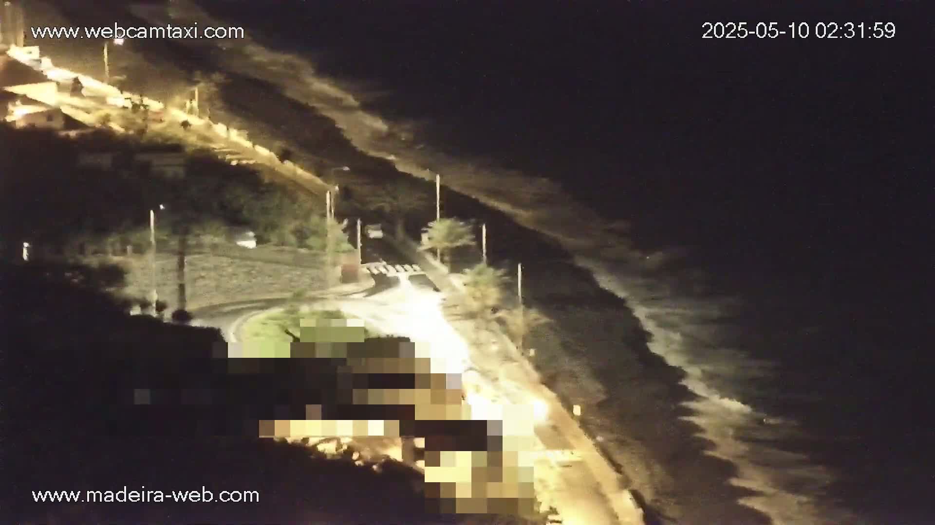 At night, large waves crash on a shoreline near a road and buildings, with some vehicles visible on the road.