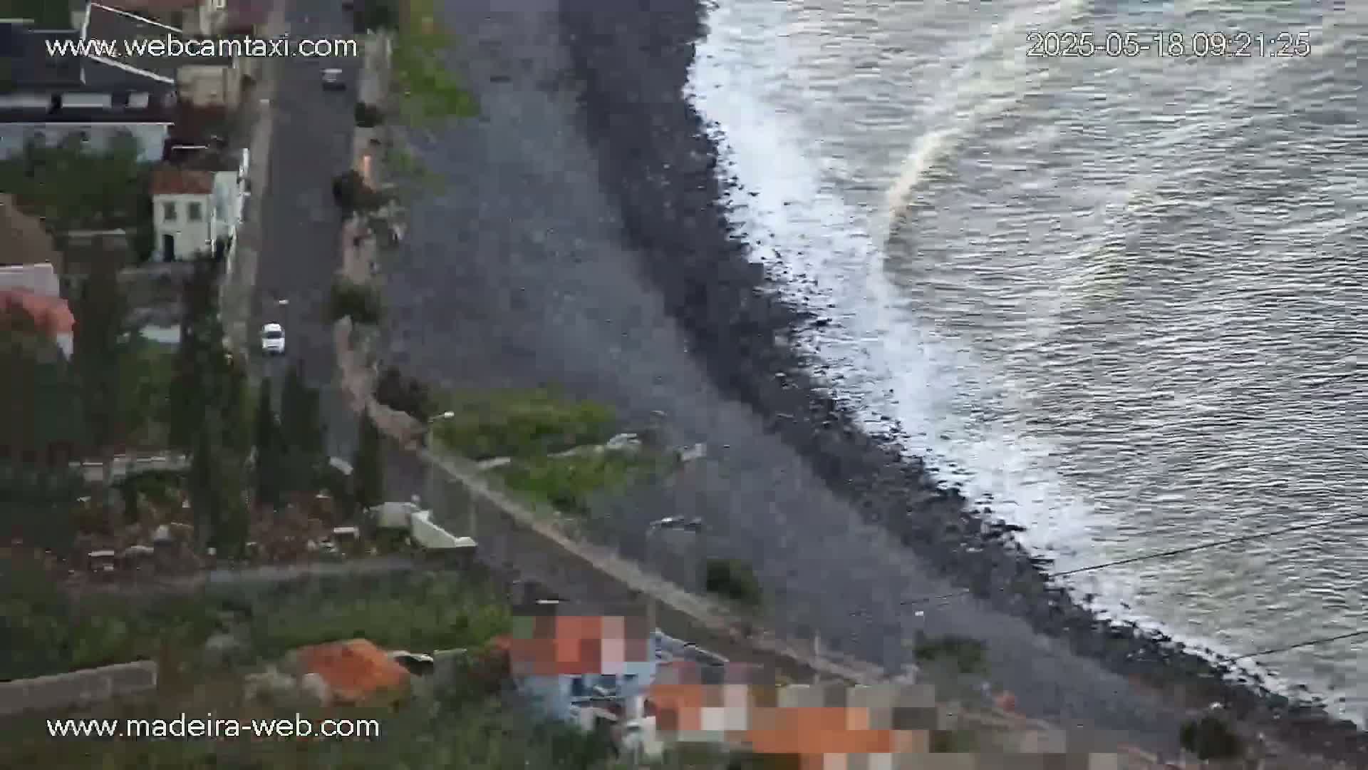 An aerial view shows a dark, rocky coastline with waves breaking on the shore next to a road and buildings.