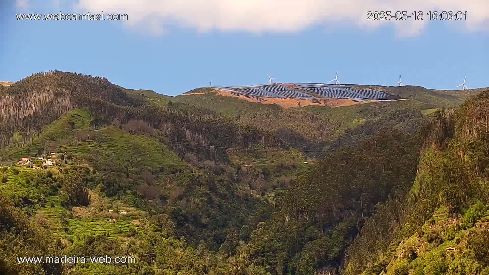 A partly cloudy sky overlooks a mountainous landscape with a large solar farm and several wind turbines on a distant hilltop, and lush green hillsides in the foreground.