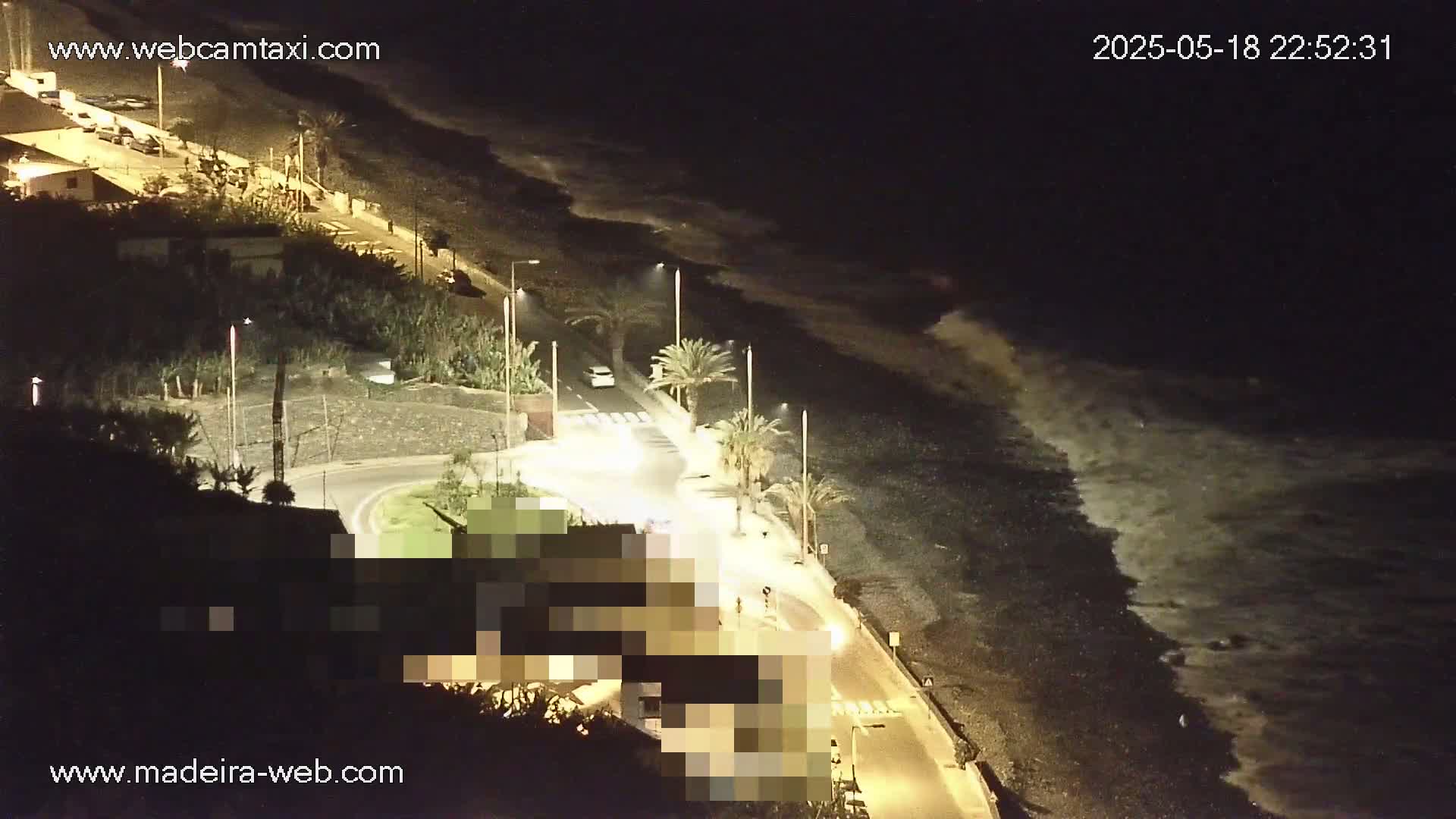 A nighttime coastal scene shows a dark, pebble beach with waves gently lapping the shore, next to a road with palm trees and buildings, illuminated by streetlights.