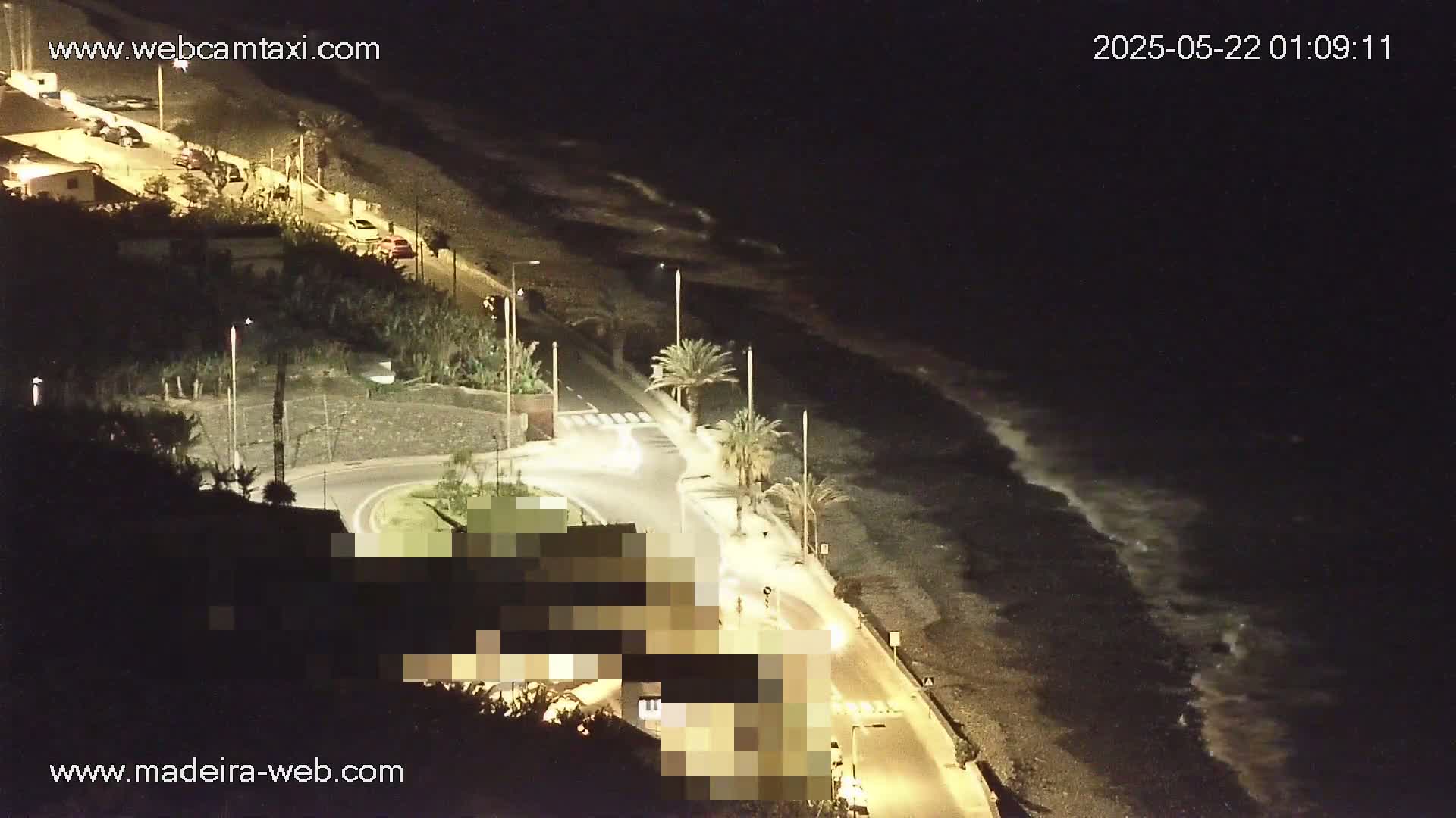 A nighttime, high-angle view shows a dark, pebble beach with gentle waves lapping the shore, alongside a roadway lined with palm trees and buildings, lit by artificial light.