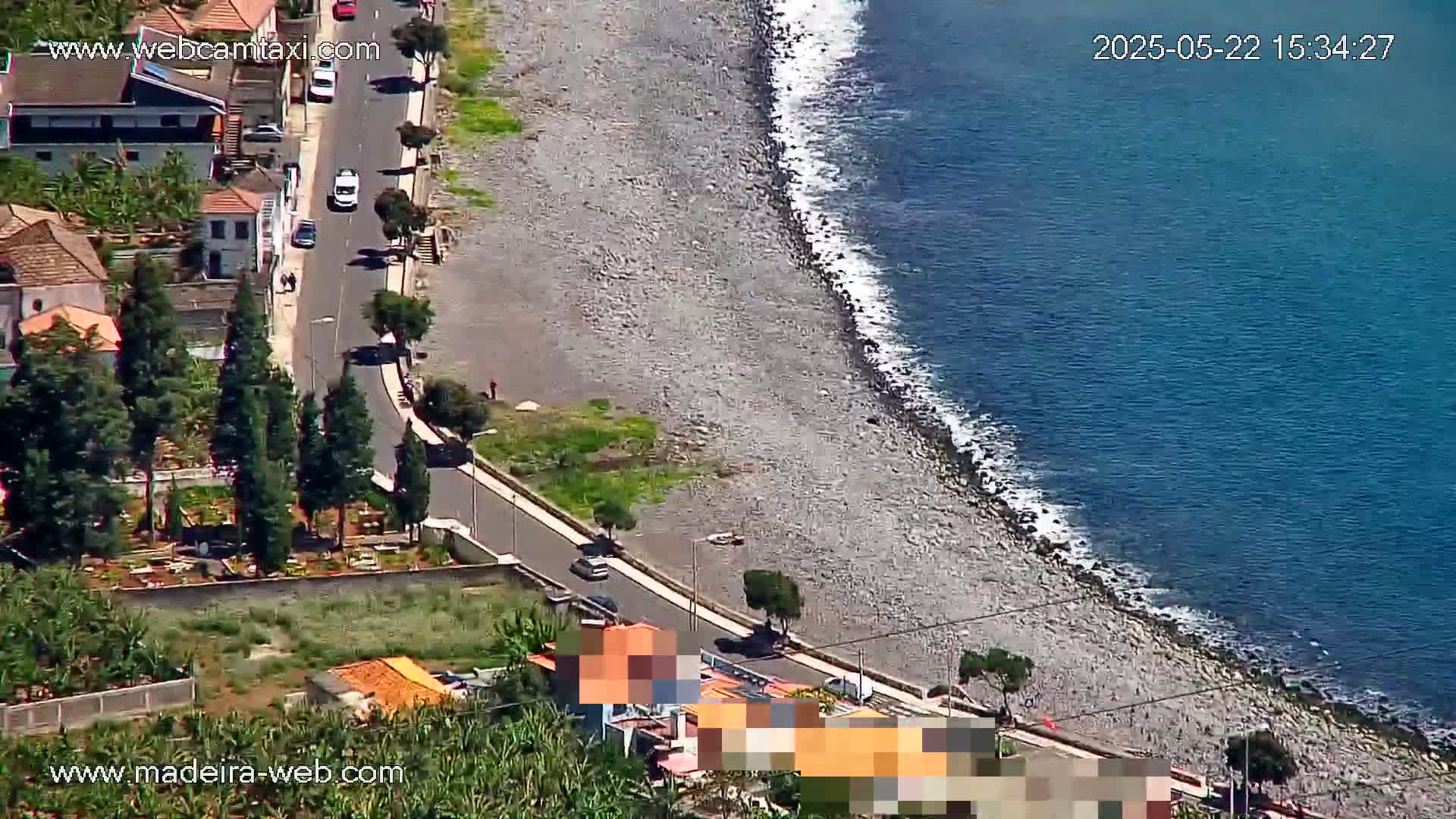 An aerial view shows a rocky beach meeting a calm blue ocean, with a road and buildings alongside the shore on a sunny day.