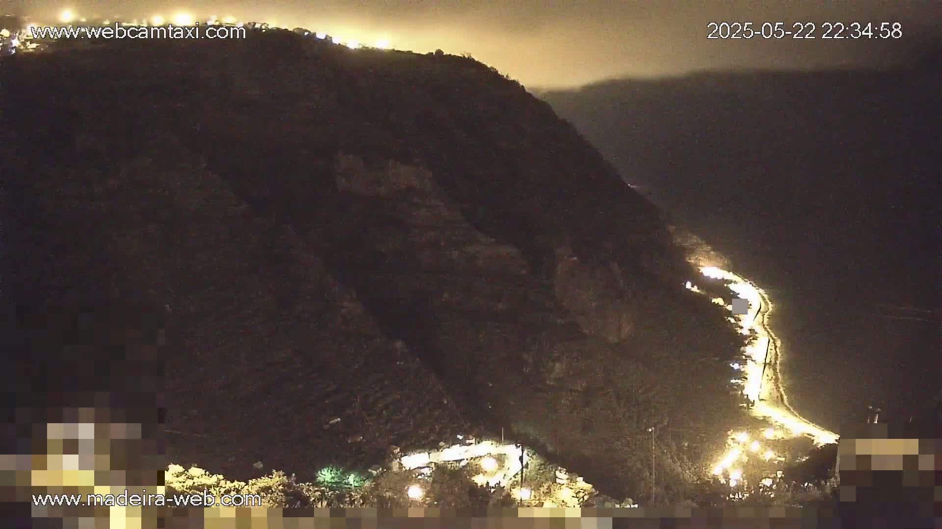 A nighttime, high-angle view shows a winding road illuminated by lights along the base of a dark mountain.