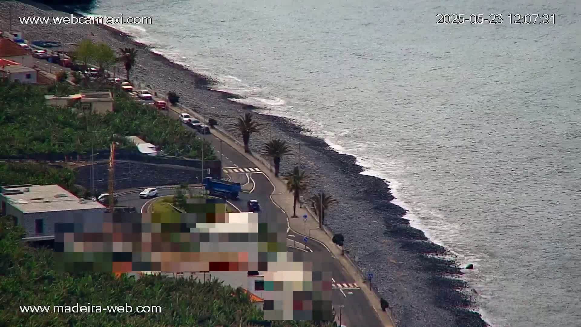 A dark-colored, rocky beach with gentle waves and a road lined with palm trees runs alongside buildings and vegetation.