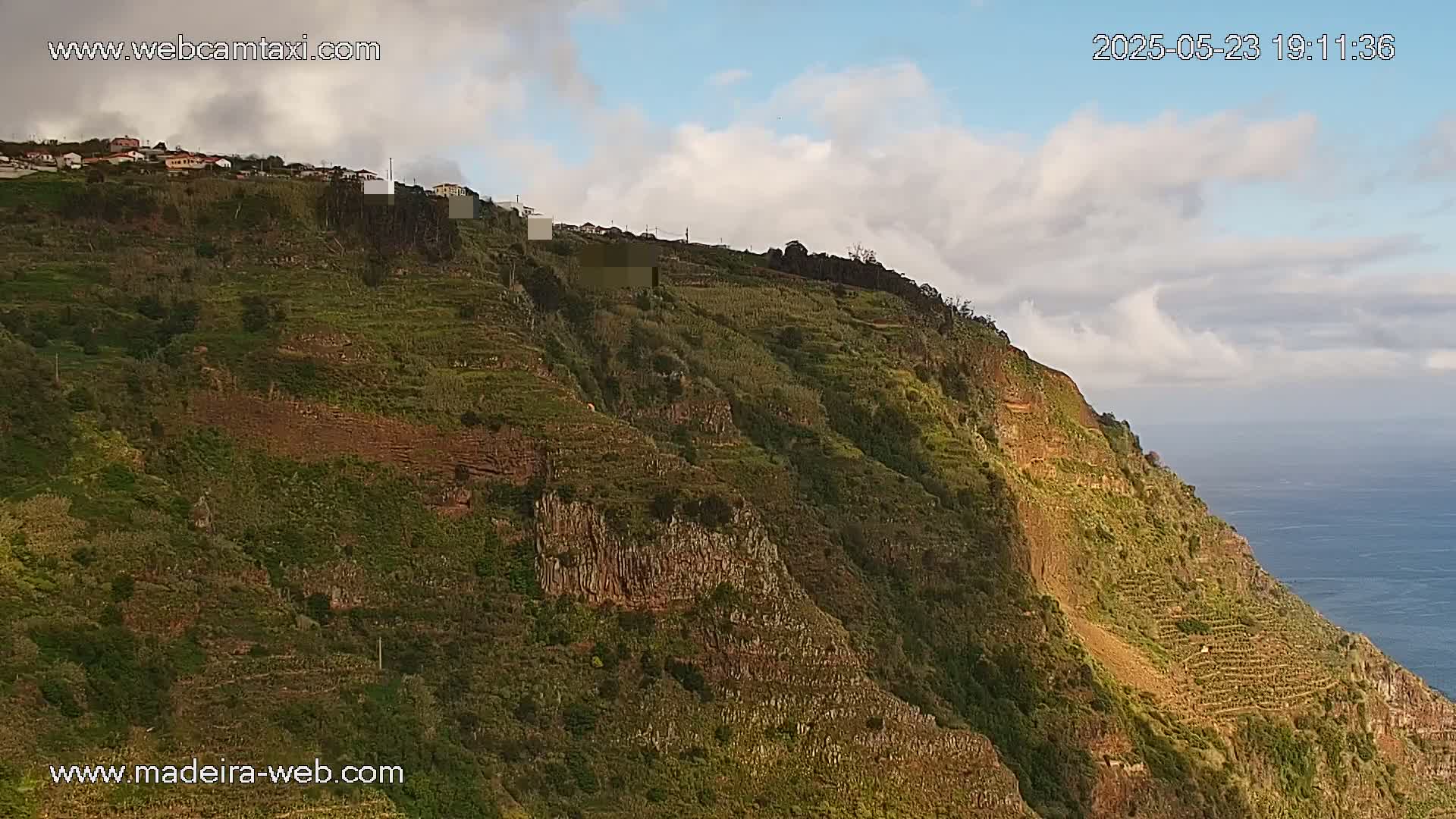 A partly cloudy sky overlooks a steep, terraced hillside with lush green vegetation and a rocky cliff face descending to a calm ocean.
