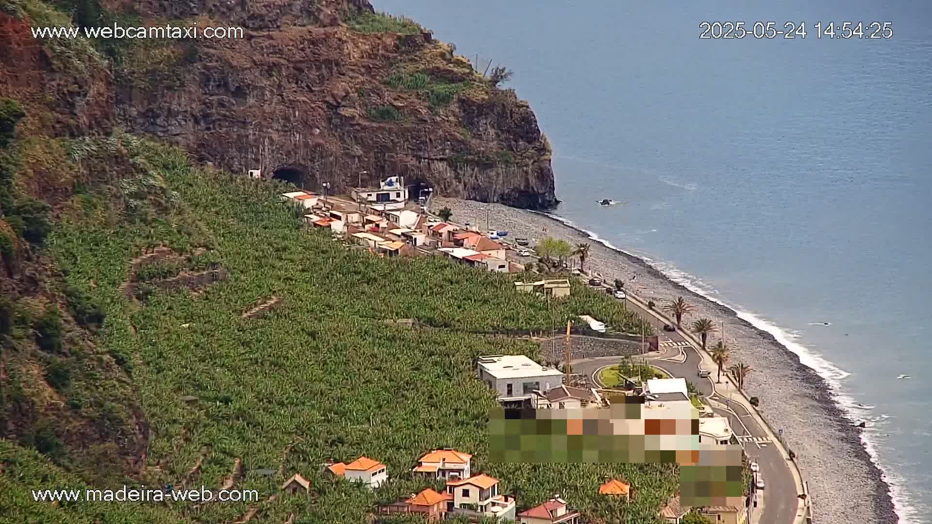 A coastal village nestled against a steep, rocky cliff overlooks a calm ocean with a pebble beach and a road curving along the shore.