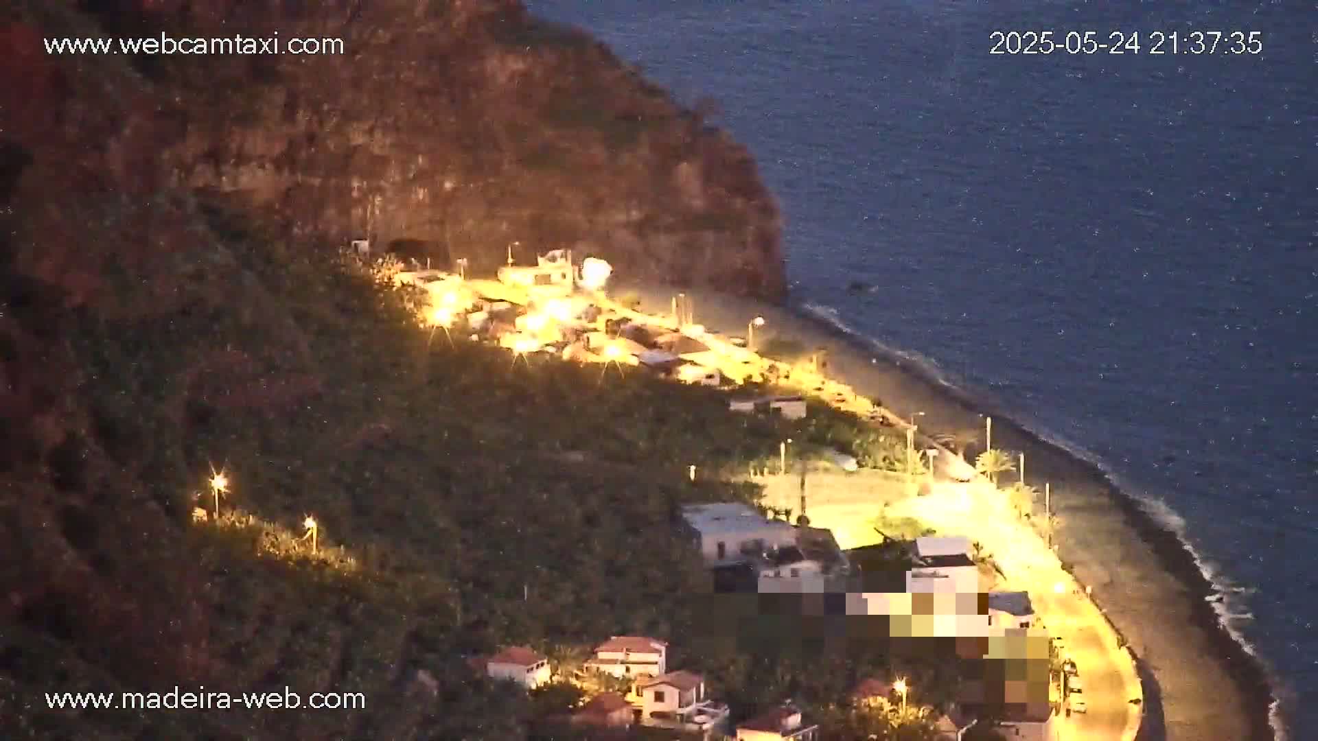 A nighttime, aerial view shows a dark-colored beach next to a cliffside village illuminated by streetlights, with a calm, dark-blue ocean in the background.