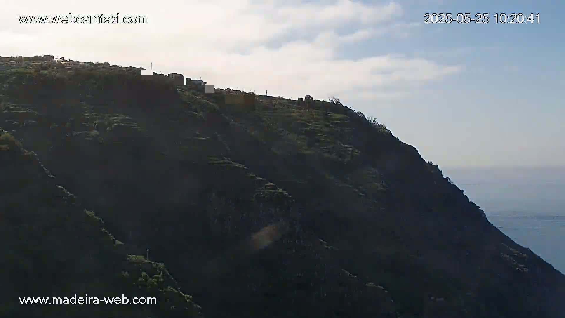 A hillside village is perched atop a dark, steep cliff overlooking a hazy ocean under a partly cloudy sky.