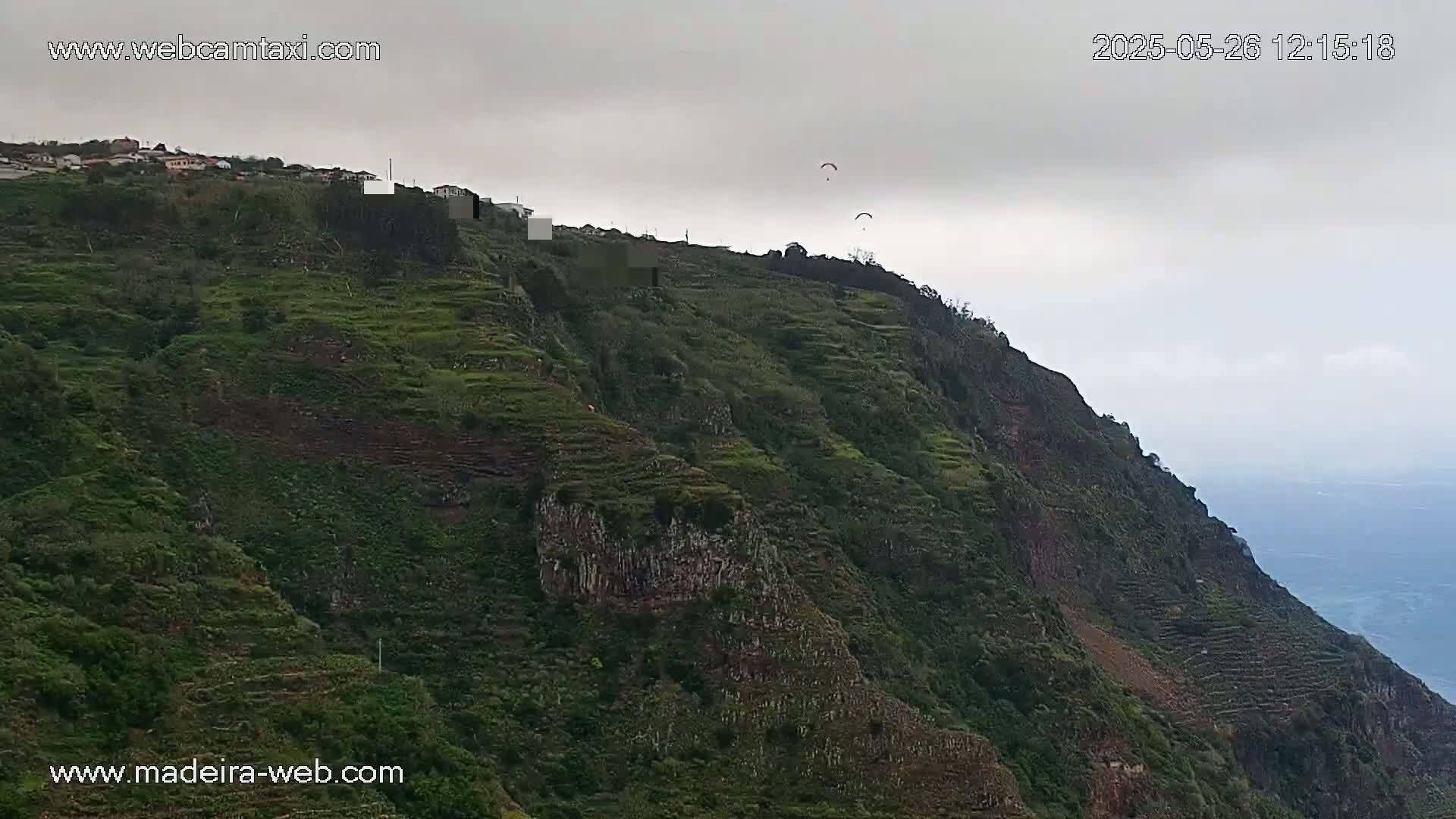 A steep, terraced hillside covered in lush green vegetation under an overcast sky, with several paragliders visible in the distance.