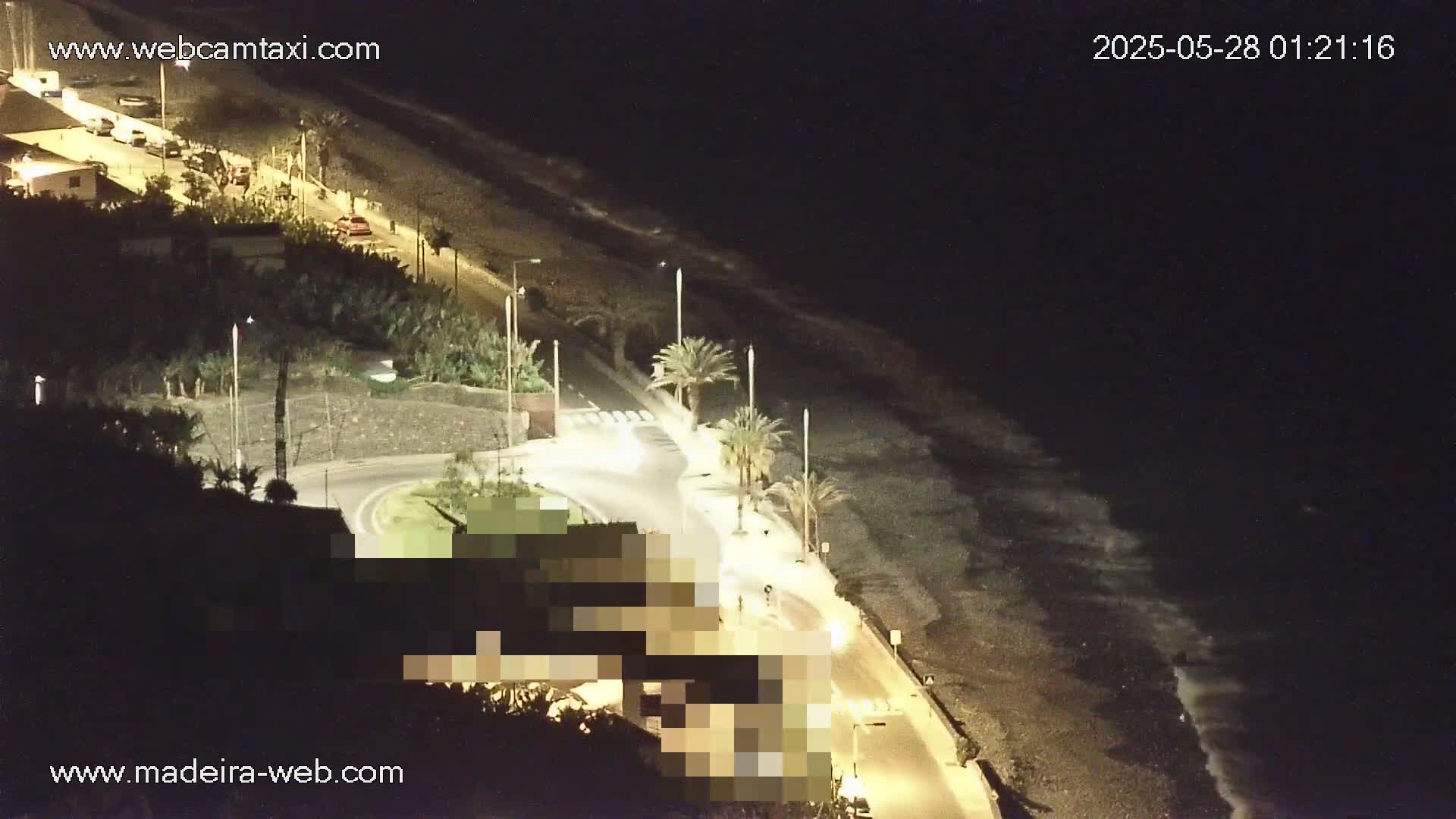 A nighttime, high-angle view shows a dark, pebble beach with gentle waves lapping the shore next to a road with sparse traffic and palm trees, all illuminated by artificial light.