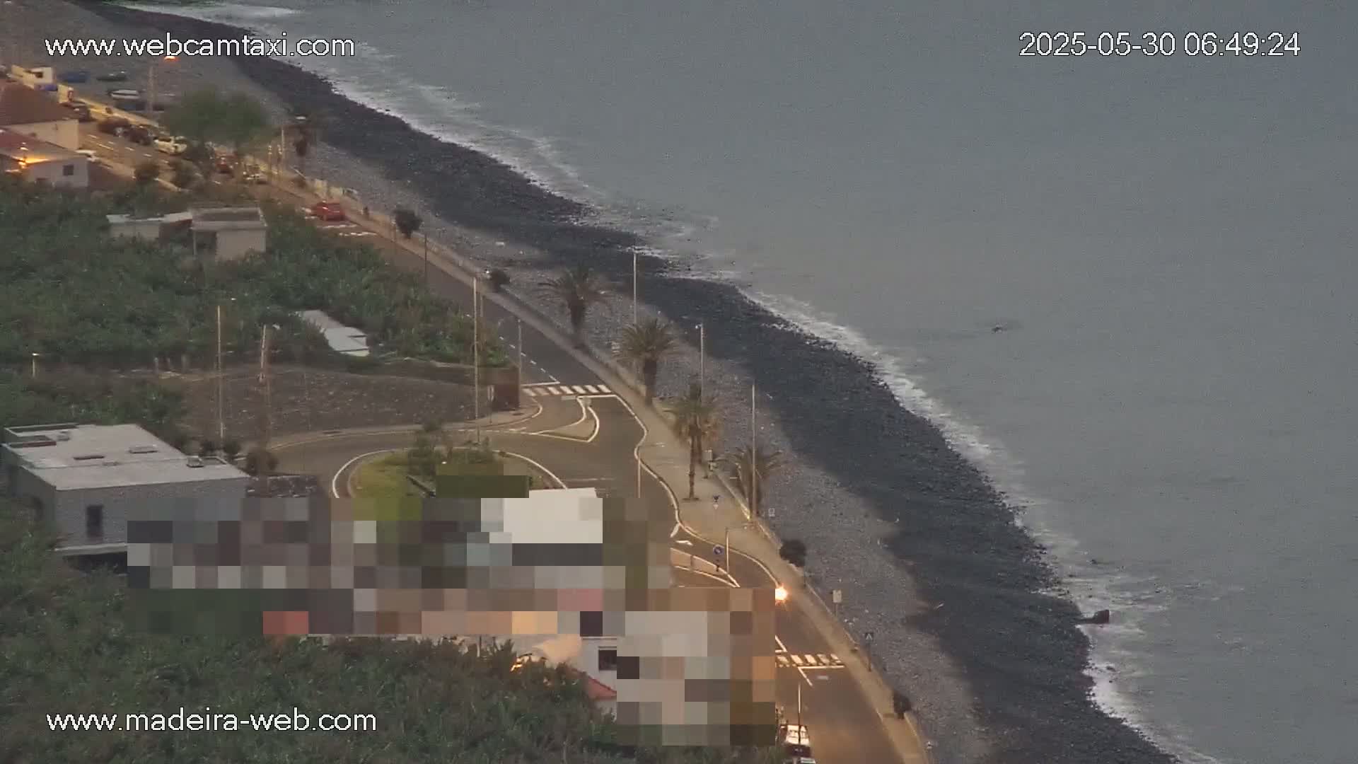 A dark, pebble beach with gentle waves under an overcast sky is bordered by a road lined with palm trees and buildings.