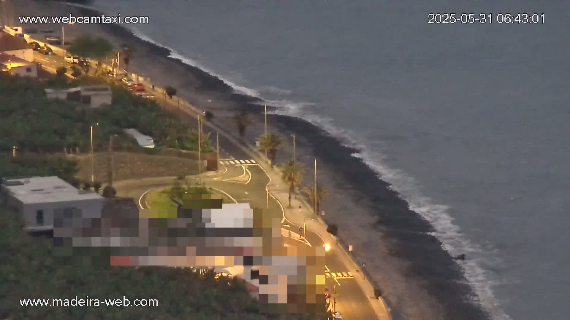 A coastal road curves around a dark-sand beach with gentle waves under a dusky sky.