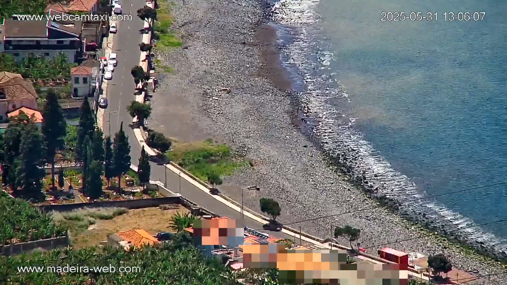 An aerial view shows a rocky beach meeting a calm ocean under a sunny sky, with a road and houses alongside the shore.