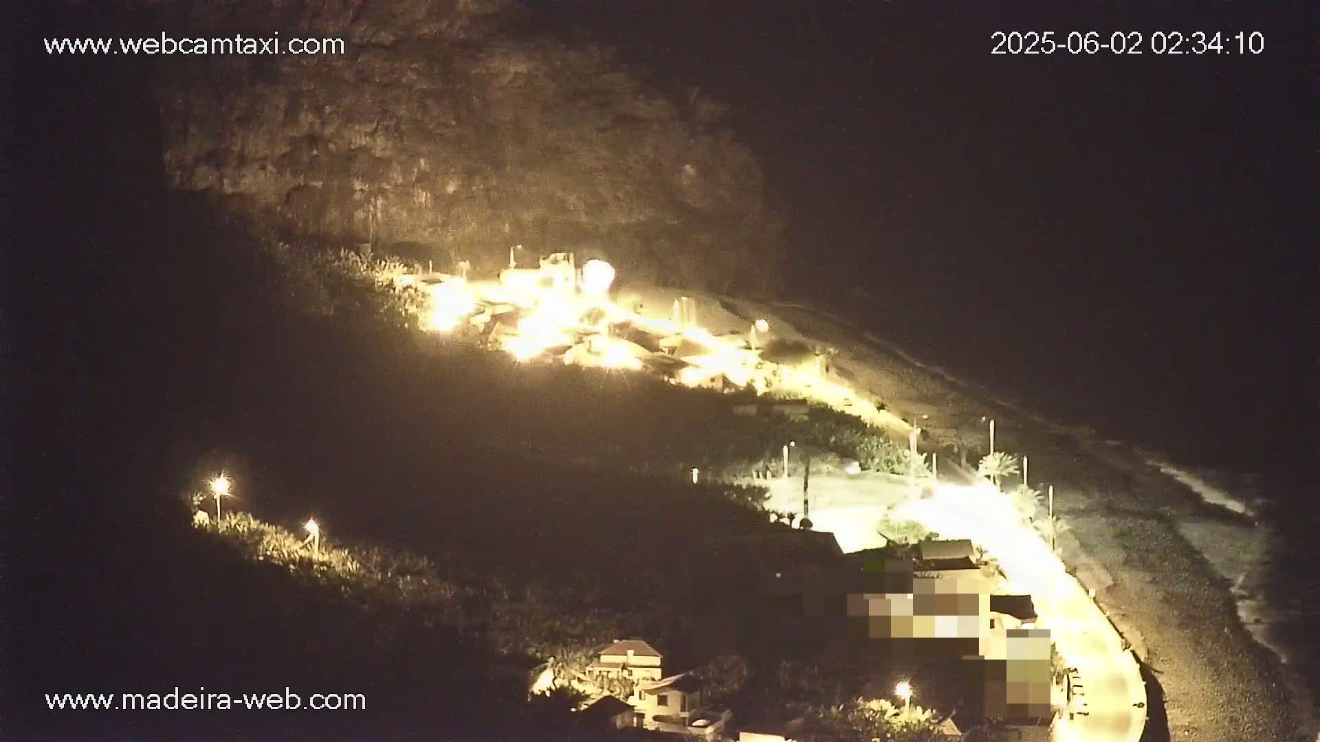 A nighttime, high-angle view shows a coastal area with buildings illuminated along a dark beach under a clear sky.