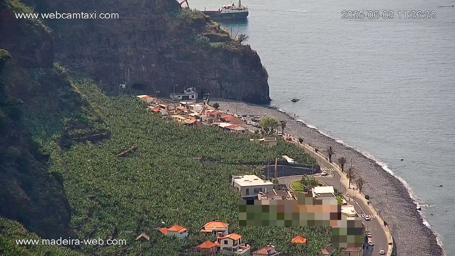 A coastal scene on a sunny day shows a rocky beach, banana plantations, and a small village nestled against a steep cliffside with a cargo ship visible offshore.
