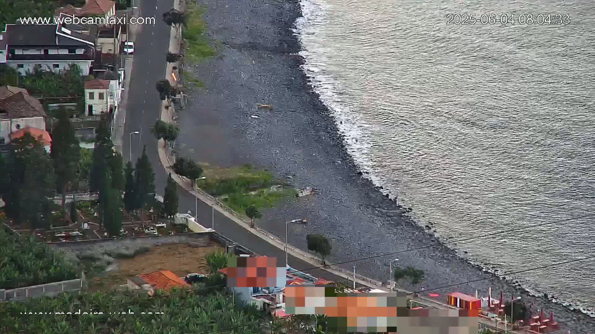 A dark-grey, pebble beach meets a calm, grey-blue ocean next to a road with buildings and vegetation on an overcast day.