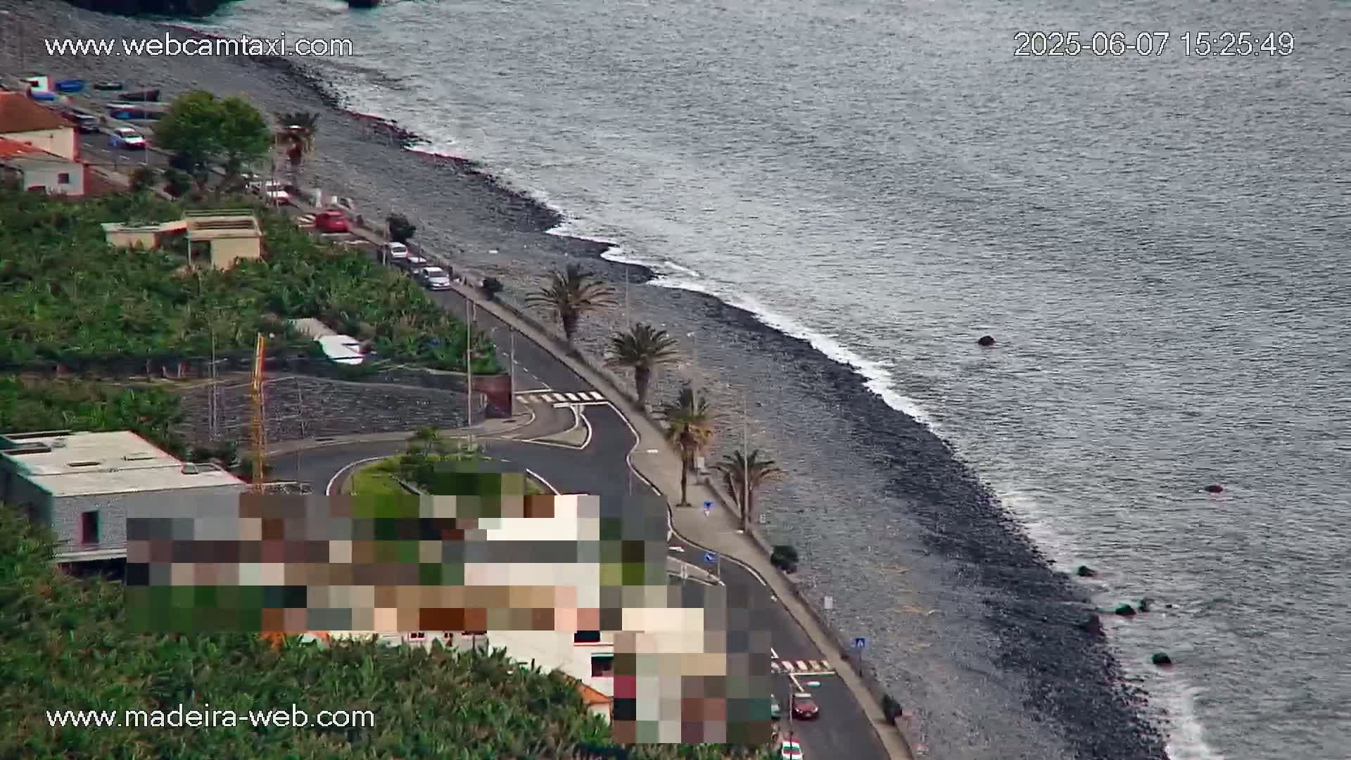 An aerial view shows a dark-grey pebble beach meeting a dark-grey sea with small waves, a coastal road lined with palm trees, and buildings nestled among green vegetation.