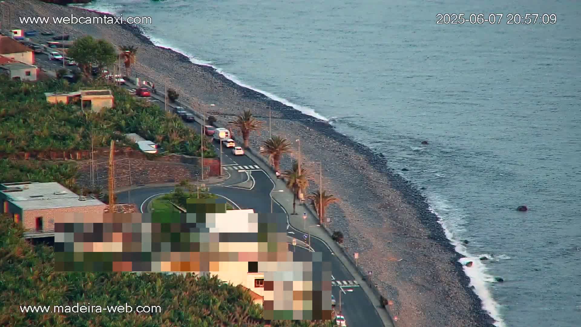 A coastal road curves along a pebble beach next to a calm ocean at dusk, with palm trees lining the roadside and several cars parked.