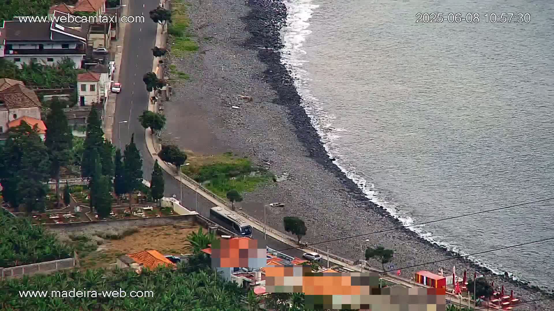 An aerial view shows a dark-grey pebble beach meeting a calm, grey-blue ocean, with a road and buildings visible inland.
