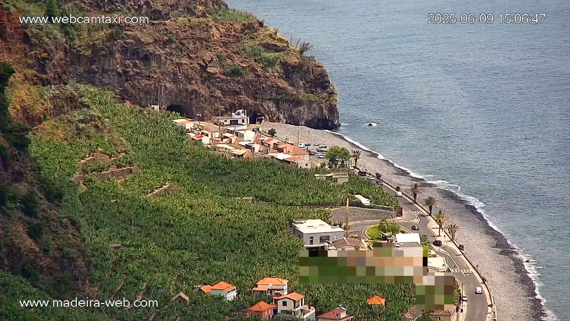 A coastal village nestled between a steep, rocky cliff and a calm ocean, with banana plantations covering the hillside.