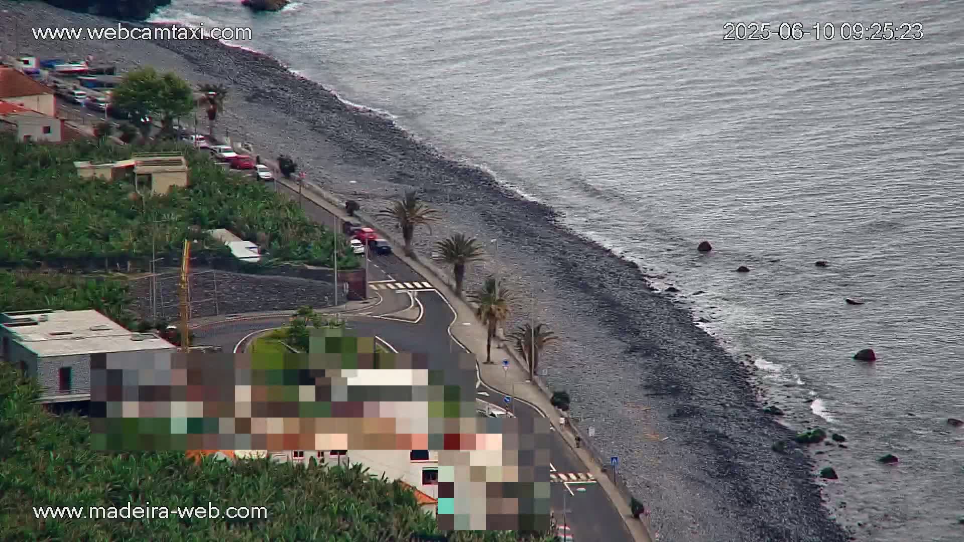 A dark-grey pebble beach borders a calm, grey ocean; a road curves along the shore past buildings and tropical plants.