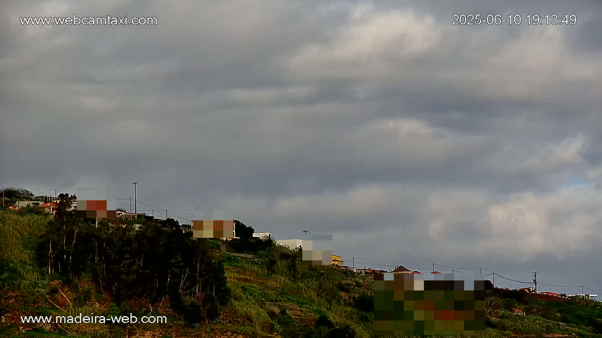 A hillside covered in dark green vegetation and houses under a mostly cloudy sky.