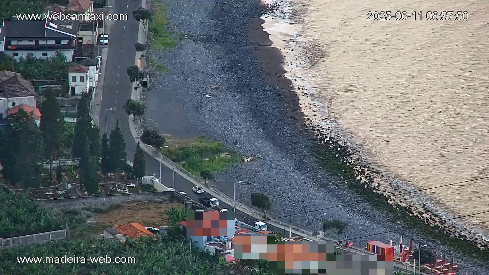 An aerial view shows a rocky coastline with gentle waves lapping the shore next to a road with several vehicles parked along it and buildings nearby.