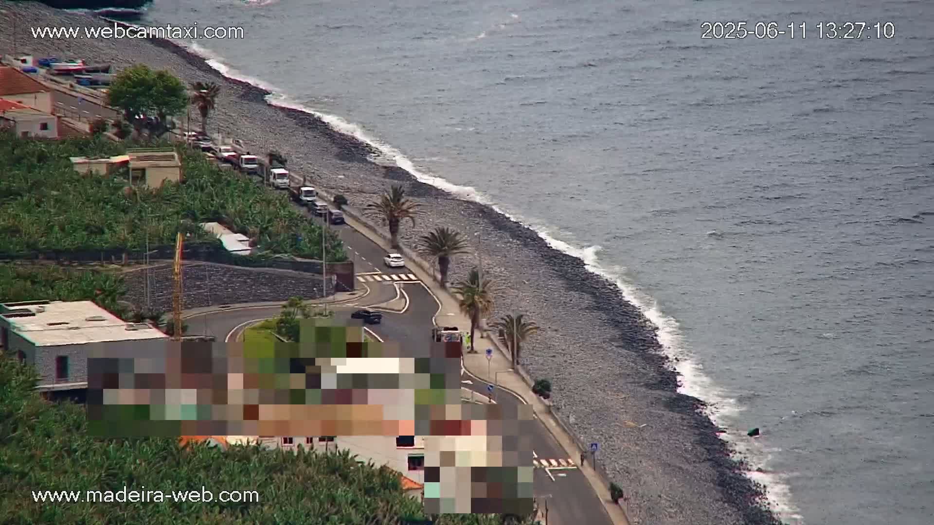 An aerial view shows a rocky coastline with gentle waves, a road lined with palm trees and parked cars, and buildings nestled among vegetation.