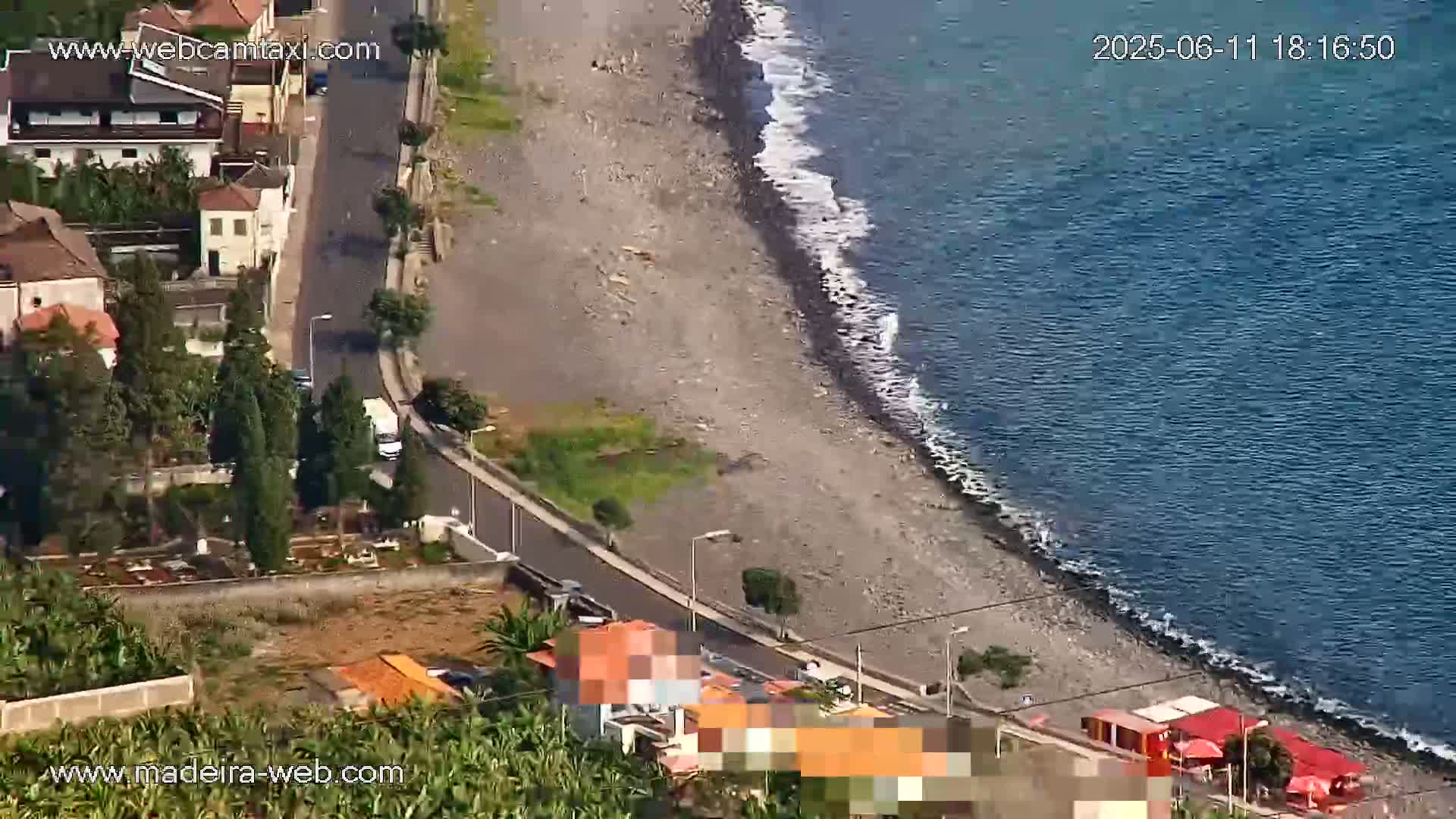 An aerial view shows a rocky beach meeting a calm ocean, with a road and buildings situated above it.