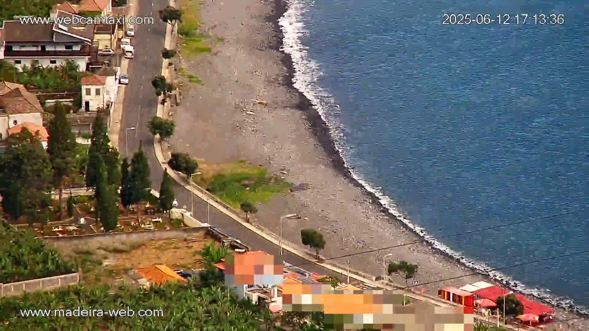 An aerial view shows a dark-colored, pebbled beach meeting a calm, dark-blue ocean, next to a road winding through a residential area with lush green vegetation.