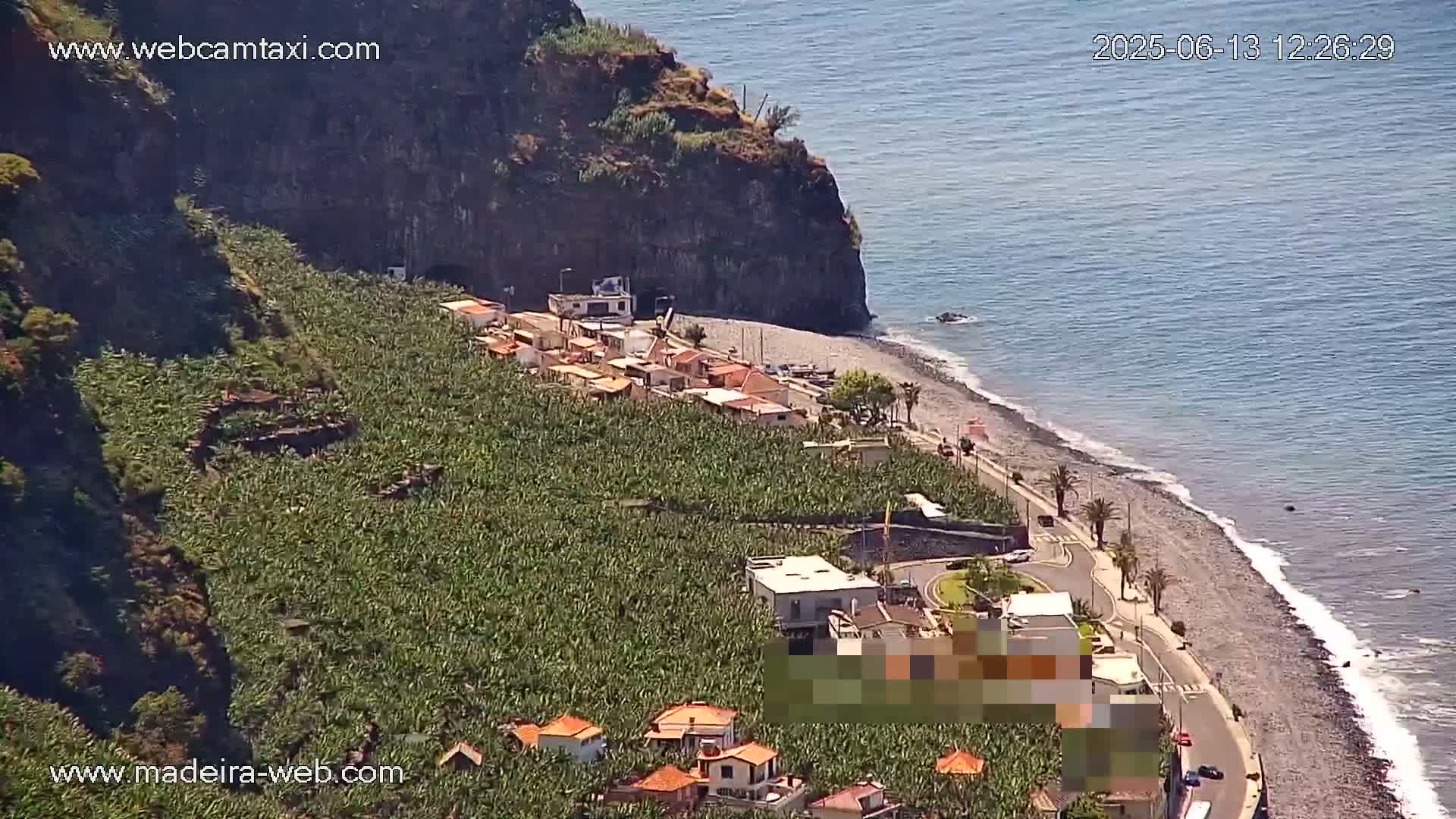 A coastal village nestled between a steep, rocky cliff and a dark-sand beach on a sunny day, with a large banana plantation covering the hillside.