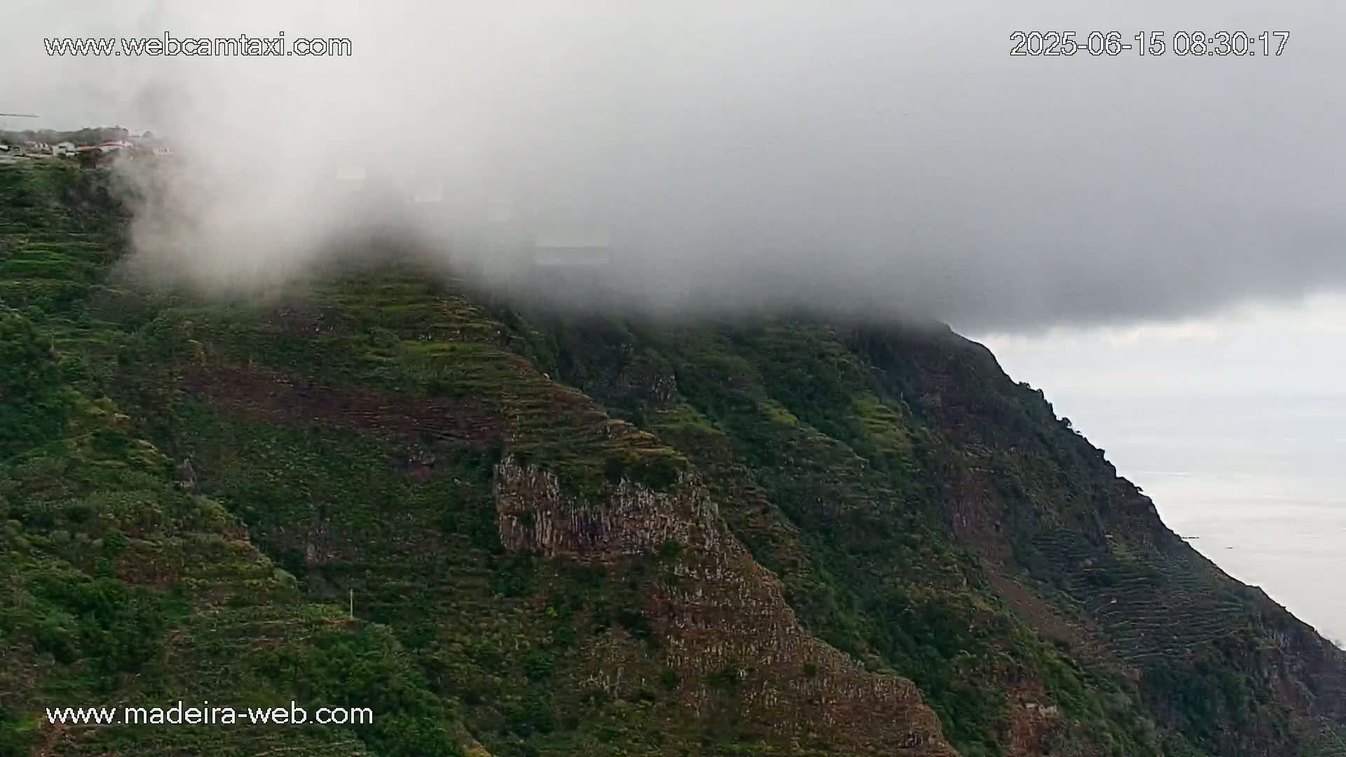 A foggy, mountainous landscape shows terraced hillsides covered in green vegetation, descending toward a calm body of water.