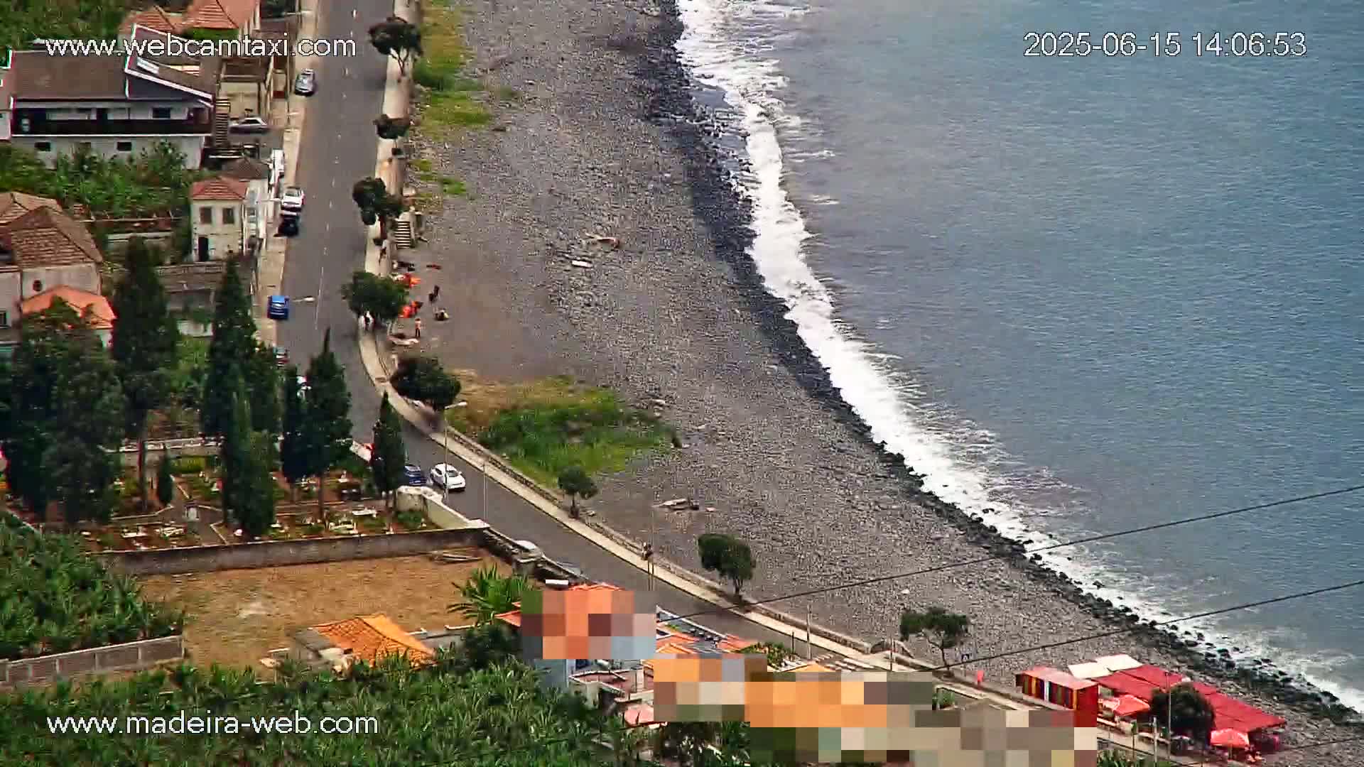 A rocky, pebble beach with gentle waves laps against the shore next to a road lined with buildings and a small cemetery, under a partly sunny sky.