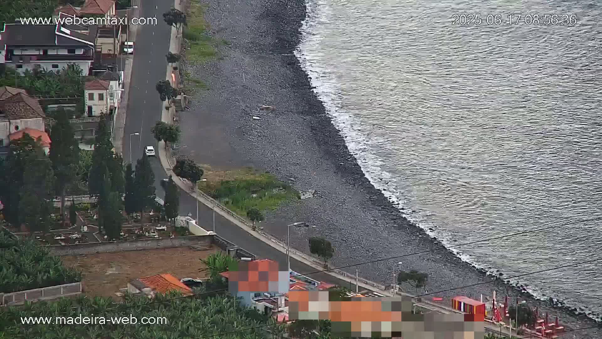 A dark, rocky shoreline meets a gray ocean with small waves, overlooking a coastal road with buildings and a cemetery.